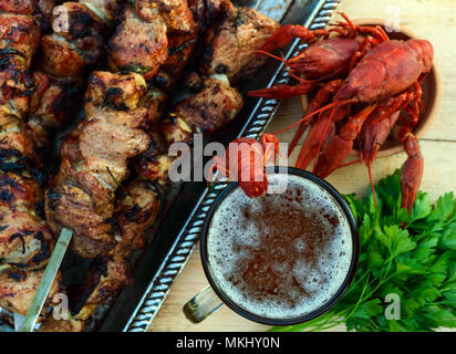 Schäumendem Bier im Glas und gekochte Languste closeup, gegrilltes Fleisch am Spieß im Hintergrund. Die Ansicht von oben. Für die Feiertage, im Freien genießen. Stockfoto