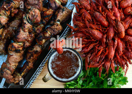 Schäumendem Bier im Glas und gekochte Languste closeup, gegrilltes Fleisch am Spieß im Hintergrund. Die Ansicht von oben. Für die Feiertage, im Freien genießen. Stockfoto