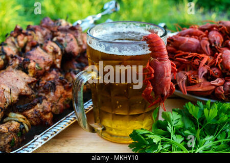 Schäumendem Bier im Glas und gekochte Languste, gegrilltes Fleisch am Spieß im Hintergrund. Für die Feiertage, im Freien genießen. Stockfoto