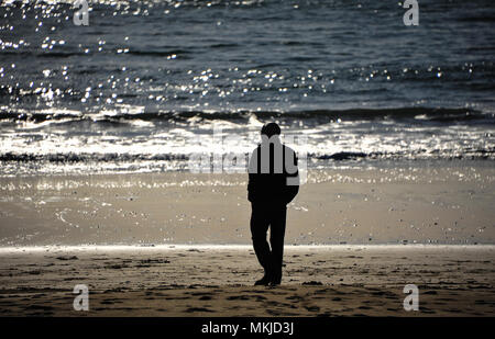 Ein Mann steht gerade Wellen brechen entlang der Küste, wie die Sonne über eine wunderschöne, silberne Landschaft setzt. Beach Szene, bis Silhouette der Person schliessen Stockfoto