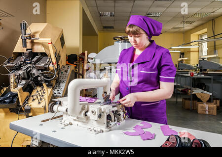 Eine junge Frau arbeitet als Näherin in einer lila Unifrome ist echt Leder Kinder Schuhe auf eine Nähmaschine näht, im Hintergrund eine Nähmaschine arbeiten Stockfoto