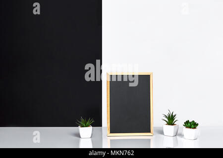 Holzrahmen mit schwarzen Platz für Text. Mock up. Stilvolle Inneneinrichtung. Grüne Pflanze in einem weißen Topf auf Schwarz-weiße Wand im Hintergrund Stockfoto