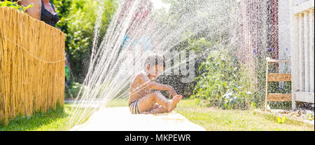 Junge Abkühlung mit Gartenschlauch, Familie im Hintergrund Stockfoto