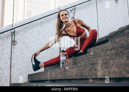 Passen junge Frau sitzt auf der Treppe im Freien und Strecken die Beine. Läuferin ihr Bein Muskeln lösen in den Morgen. Stockfoto