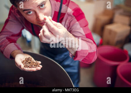 Bild des jungen Mannes mit Kaffeebohnen in der Hand neben Röster Stockfoto