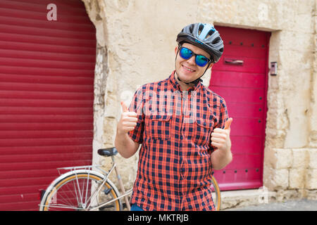 Stattliche männliche Radfahrer geben ein "Daumen hoch" auf rotem Hintergrund im Freien Stockfoto