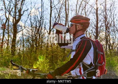 Ein Radfahrer in Sportswear ist Trinkwasser aus einer Flasche in den Wald an einem sonnigen Tag. Stockfoto