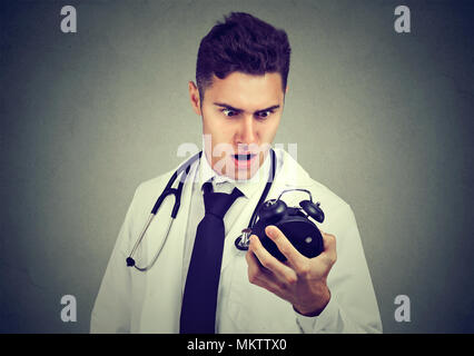 Junge Mann in der medizinischen Outfit mit der Zeit Druck auf die Uhr auf grauem Hintergrund suchen Angst. Stockfoto