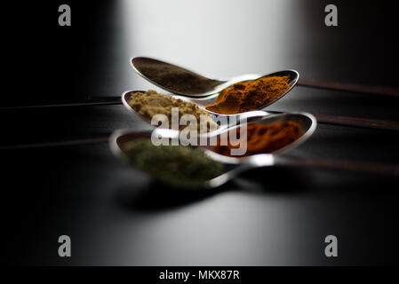 Gewürze farbige in Silber Löffel, auf einem schwarzen Holztisch gelegt. Pfeffer, Pfeffer, Salz, Basilikum, Senf Stockfoto