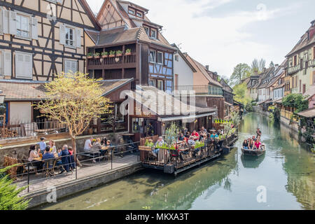 Gäste genießen Sie einen warmen Frühlingstag in einem Café am Kanal in Colmar Frankreich während andere Schwimmer durch auf einem traditionellen Barke. Stockfoto