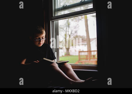 Ein 11-jähriger Junge sitzt in einem offenen Fenster und liest ein Buch. Stockfoto