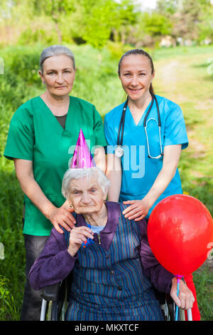 Ältere behinderte Frau in einem Rollstuhl feiert Geburtstag mit freundlichen weiblichen Pflegepersonen im Freien Stockfoto