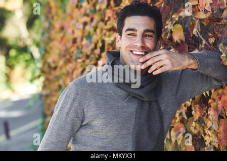 Stattlicher Mann mit blauen Augen tragen Winter Kleidung im Herbst Blätter Hintergrund lächelnd. Junge männliche mit swater und Schal. Stockfoto