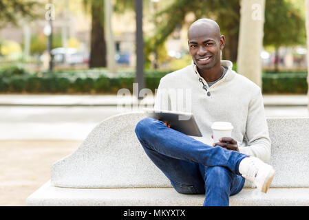 Schwarze jungen Mann mit Tablet Computer und Kaffee im städtischen Hintergrund. Junge afrikanische Kerl mit rasiertem Kopf legere Kleidung. Stockfoto