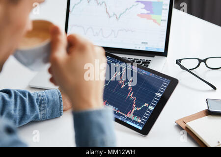 Junge Unternehmer sitzt zu Hause am Tisch, Arbeiten auf Tablet mit Grafiken, Diagrammen auf Monitor. Crypto Währungen, Trader mit Tablet-PC und Computer. Stockfoto