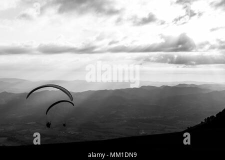 Einige Gleitschirme fliegen über eine Berglandschaft, mit einigen schwachen Sonnenstrahlen im Hintergrund Stockfoto