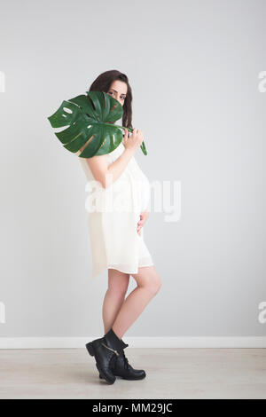 Brunette schwangere Frau tragen weiße Kleid Holding monstera Blatt Stockfoto