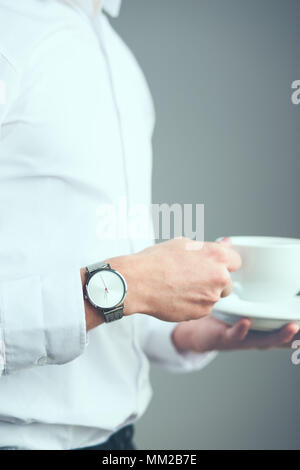 Junger Mann mit silber Armbanduhr und weiß plain T-Shirt, Tasse Kaffee. Vor plain graue Wand Stockfoto