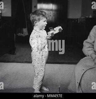 1960 s, historischen, einen Jungen in seinem Schlafanzug steht auf dem Teppichboden der Schlafzimmer seiner Eltern, eines seiner neuen Geburtstagsgeschenke, ein Spielzeugauto, England, UK.