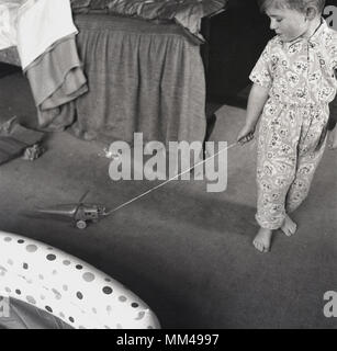1960 s, historischen, einen Jungen in seinem Schlafanzug sein neues Spielzeug Flugzeug am Boden entlang seiner Eltern Schlafzimmer zieht, nachdem es gerade als Geburtstagsgeschenk, England, UK. Stockfoto