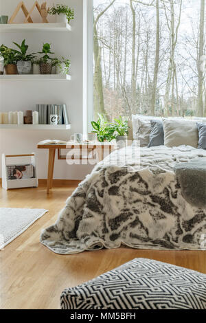 Gemusterte Puff in der Nähe von Bed mit Fell in der gemütlichen Schlafzimmer Einrichtung mit Pflanzen und mit Blick auf den Wald Stockfoto