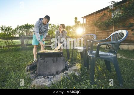 Im Sommer auf der Gartenterrasse. Junges Paar Grillen gegen Chalet am Sonnenuntergang. Stockfoto