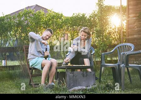 Im Sommer auf der Gartenterrasse. Junges Paar Grillen gegen Chalet am Sonnenuntergang. Stockfoto