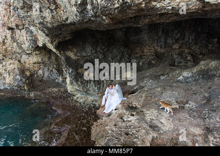 Paar Brautpaar Braut und Bräutigam Lachen und Lächeln zu einander, glücklichen und freudigen Moment. Der Mann und die Frau in der Hochzeit Kleidung sitzen auf dem Rock Hintergrund. Stockfoto