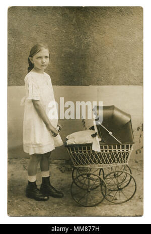 Foto im frühen 20. Jahrhundert, ein schönes Mädchen in einem weißen Kleid und schwarzen Stiefeln mit einer Puppe Kinderwagen auf der Straße gegen die Wand. Stockfoto
