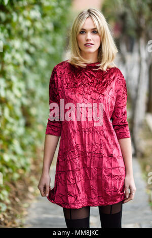 Schöne blonde Frau in grün Blätter Hintergrund. Junge Mädchen mit roten Kleid auf der Straße stand. Hübsche Frau mit krauses Haar und blaue e Stockfoto