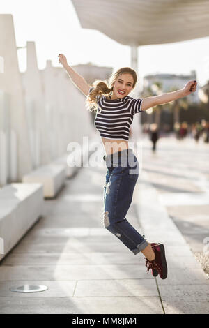 Lustige blonde Frau im städtischen Hintergrund springen. Schöne junge Mädchen tragen gestreifte T-Shirt und Blue Jeans in der Straße. Hübsche Russische Frauen mit Stockfoto