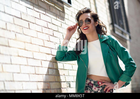 Junge brünette Frau mit Aviator Sonnenbrille. Lächelnde Mädchen, Modell der Mode, das Tragen von grünen moderne Jacke, stehen im städtischen Hintergrund. Stockfoto