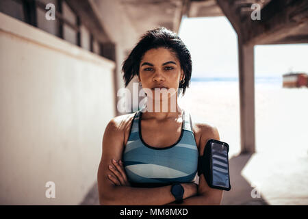 Portrait von selbstbewusste Frau Athleten stehen im Freien mit verschränkten Armen und Kamera. Mit Handy weiblichen am Armband eine Pause Stockfoto