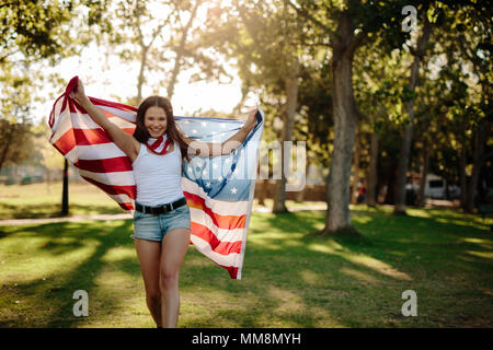 Schöne junge Frau zu Fuß in den Park und wehende amerikanische Flagge. Lächelnde Mädchen mit der amerikanischen Flagge im Park. Stockfoto