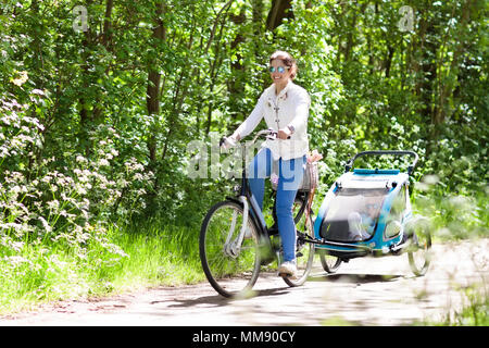 Junge Mutter mit Baby Reiten Fahrrad Fahrradanhänger im sonnigen Sommer Park. Fit aktive Frau Radfahren mit Kind. Sicherer Transport von kleinen Kindern. Mom Stockfoto