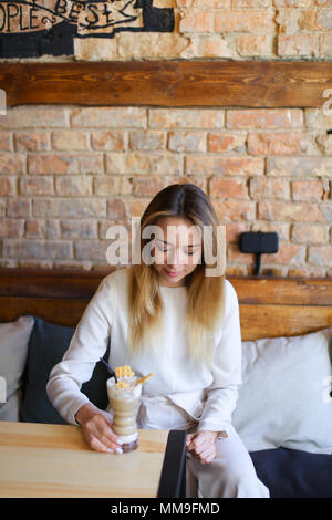 Junge Frau sitzt in der Nähe von Tabelle mit Tasse Kaffee und für Fotos posiert Stockfoto