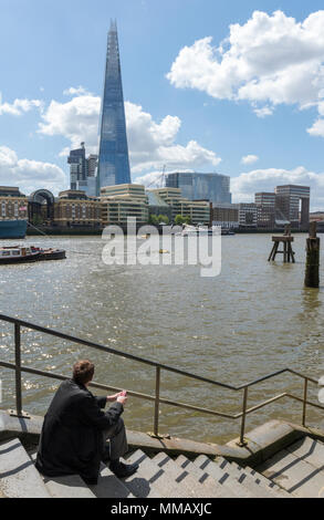 Ein Mann oder Geschäftsmann Sitzen durch die Seite der Themse im Zentrum von London, tief in Gedanken oder Denken mit dem shard Bürogebäude auf der anderen Themseseite Stockfoto