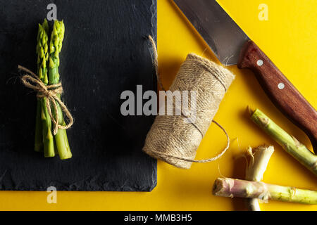 Spargel. Bündel frischer Spargel auf einer Platte von Schiefer mit einem Messer gebunden, eine Rolle von Seil und Scheiben von Spargel Schneiden auf gelben Hintergrund. Gesund Stockfoto