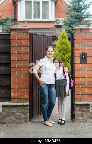 Glückliche junge Mutter mit Tochter in Schuluniform vor dem Haus posing Stockfoto