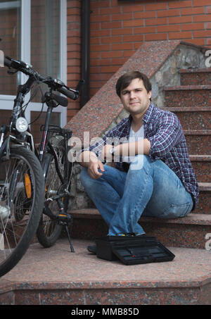 Lächelnden jungen Mann sitzt auf Haus Veranda und Reparatur Fahrrad Stockfoto