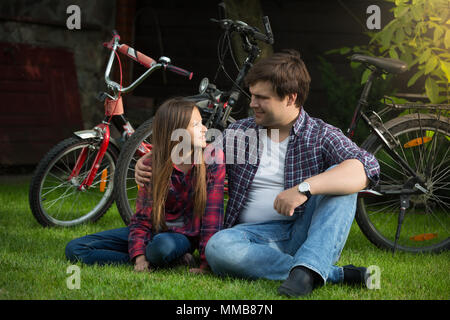 Lächelnde Mädchen ihren Vater sitzen auf dem Rasen im Park auf der Suche nach Fahrrad Stockfoto