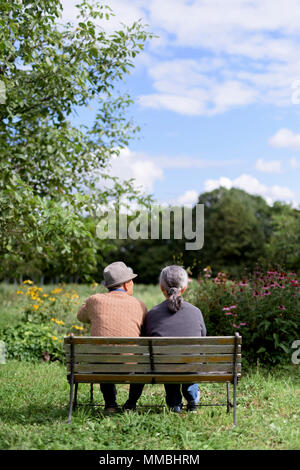 Mann und Frau, Rückansicht der älteren Mann mit Hut und eine Frau sitzen nebeneinander auf einer Bank in einem Garten. Stockfoto