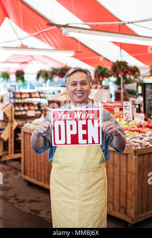 Lächelnder Mann mit Schürze zu einem Obst- und Gemüsemarkt, Holding aloft Öffnen unterzeichnen. Stockfoto