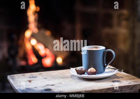 Nahaufnahme von Becher mit heißen Getränk auf hölzernen Tisch vor einem Kamin. Stockfoto