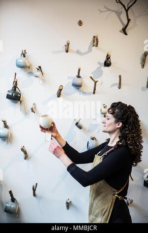 Nahaufnahme von Frau braun-gelockten Haar zu tragen Schürze steht in der Keramik shop, hängende Tassen auf kleine Zweige auf eine Wand. Stockfoto