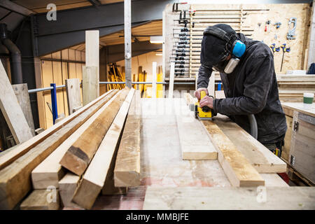 Mann, Gehörschutz, Schutzbrille und Staubmaske stehend in einem Lager, schleifen Dielen aus recyceltem Holz. Stockfoto