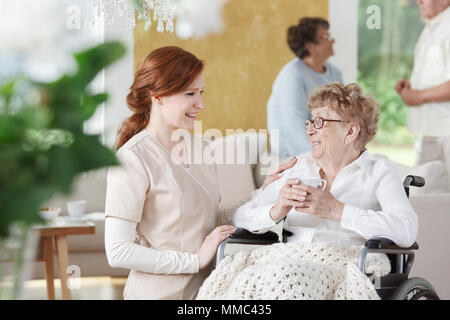 Ältere Frau sitzt im Rollstuhl Neben Krankenschwester und Halten einer Tasse Kaffee Stockfoto