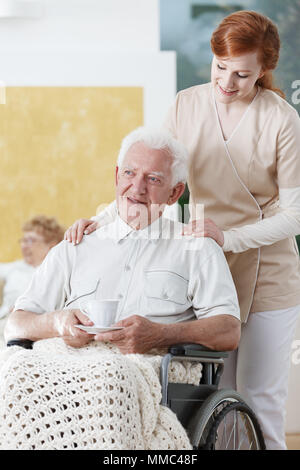 Krankenschwester steht neben behinderten älteren Menschen auf Rollstuhl, der Tasse Kaffee Stockfoto