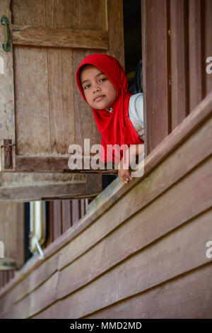 Krabi, Thailand - 2. Mai 2015: Cute muslimischen Mädchen das Tragen der roten hijap aus Holz Windows lächelnd an Besucher in ländlichen von Krabi, Thailand bewegen Stockfoto