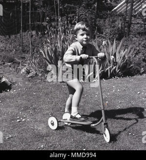 1960er Jahre, Jungen draußen auf einem Rasen und Garten stehen auf einem Dreirädrigen Roller aus Metall, England, UK. Stockfoto